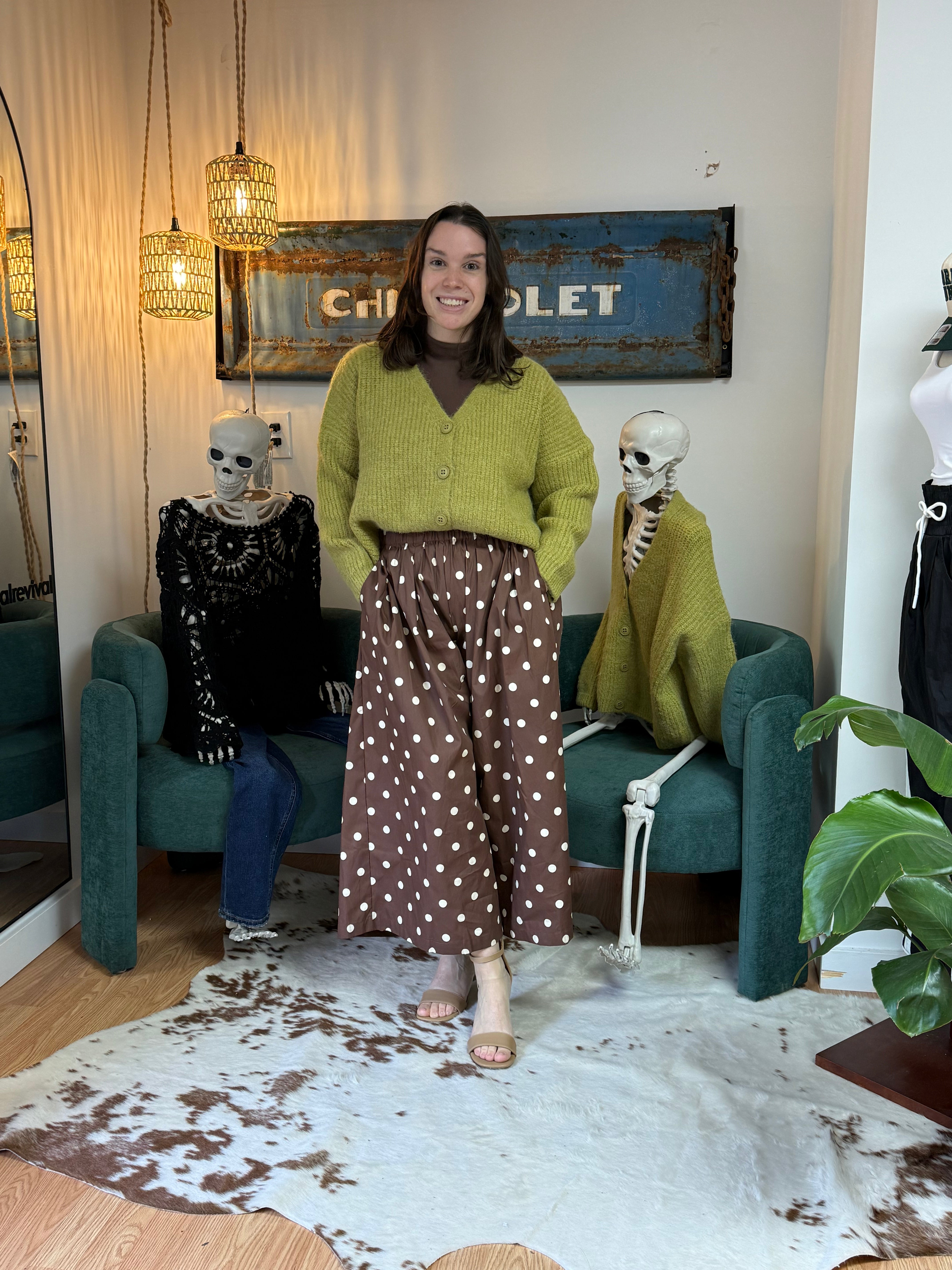 A woman stands in a styled indoor room wearing a lime green knit cardigan and a brown wide-leg skirt with white polka dots. The setting includes teal armchairs, skeleton decorations, hanging pendant lights, a rustic blue sign on the wall, and a cowhide rug on the floor.
