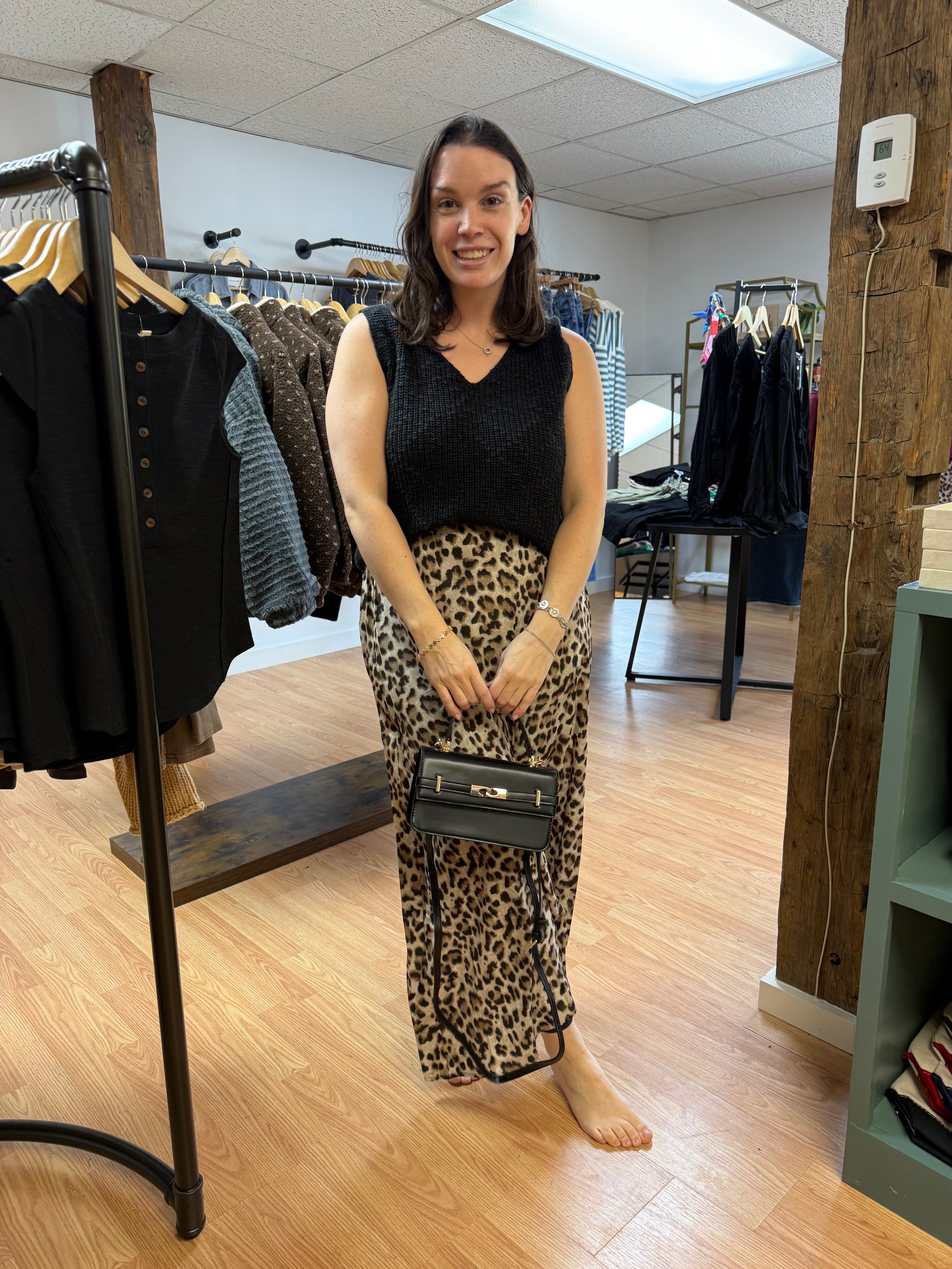 A woman stands in a clothing boutique wearing a black sleeveless top and a leopard-print maxi skirt, holding a small black handbag. Clothing racks and wooden floors are visible in the background.